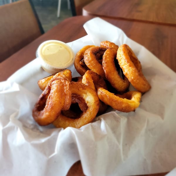 Golden-brown onion rings in a paper-lined basket with a side of dipping sauce on a wooden table.