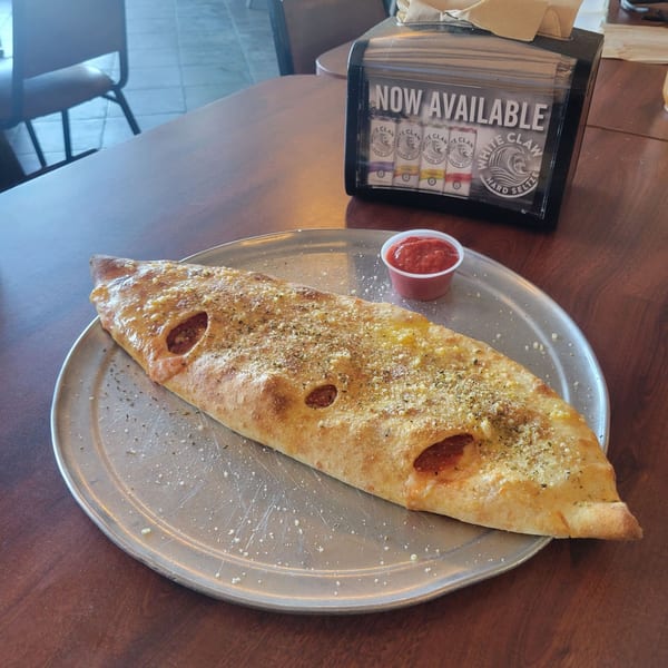A large, golden-brown calzone with pepperoni and cheese, served with marinara sauce on a metal tray.