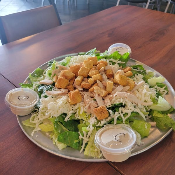 A large Caesar salad with chicken, croutons, and shredded cheese on a metal platter with dressing cups.